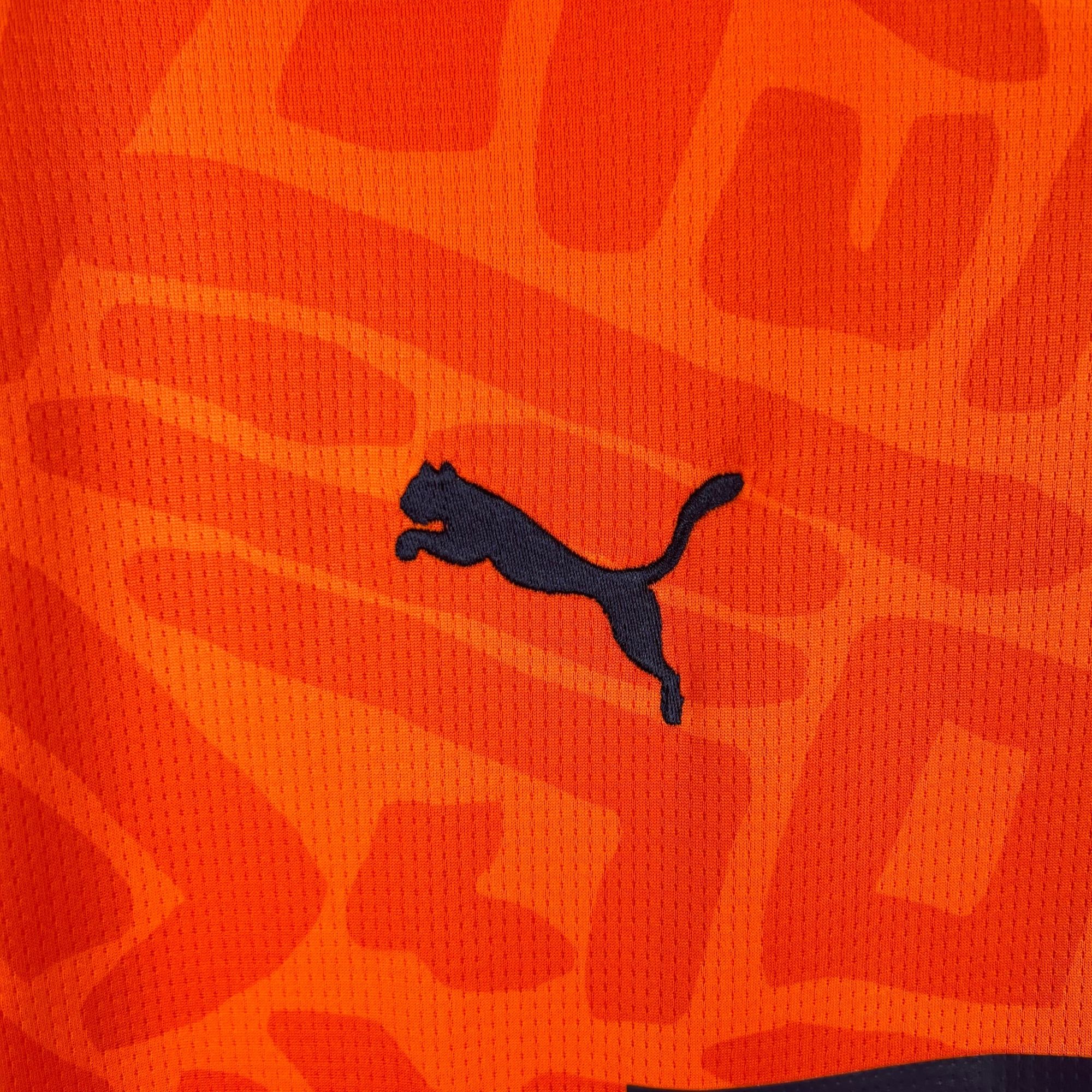 00408b5f_7fa3c68f-8bb7-4c88-a6fb-8f65678c3b29 | AE JERSEY Close-up of orange fabric with a dark blue Puma logo visible on the right edge.