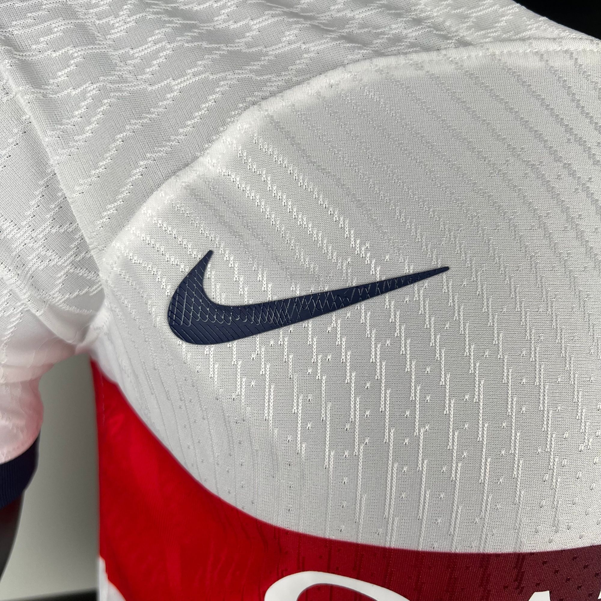 053afa21_209366fa-6ba8-4a15-b2a5-e84a6a051c7a | AE JERSEY Dark navy Nike swoosh logo on white textured fabric with a red background nearby, cropped close-up.