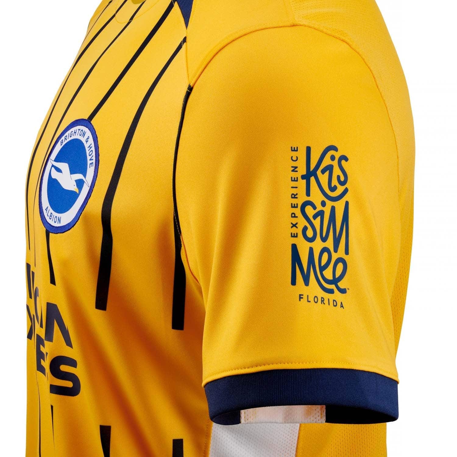 0Zn2300RpuMzQFx | AE JERSEY Close-up of a yellow soccer jersey sleeve with the Brighton & Hove Albion crest and blue sponsor text.