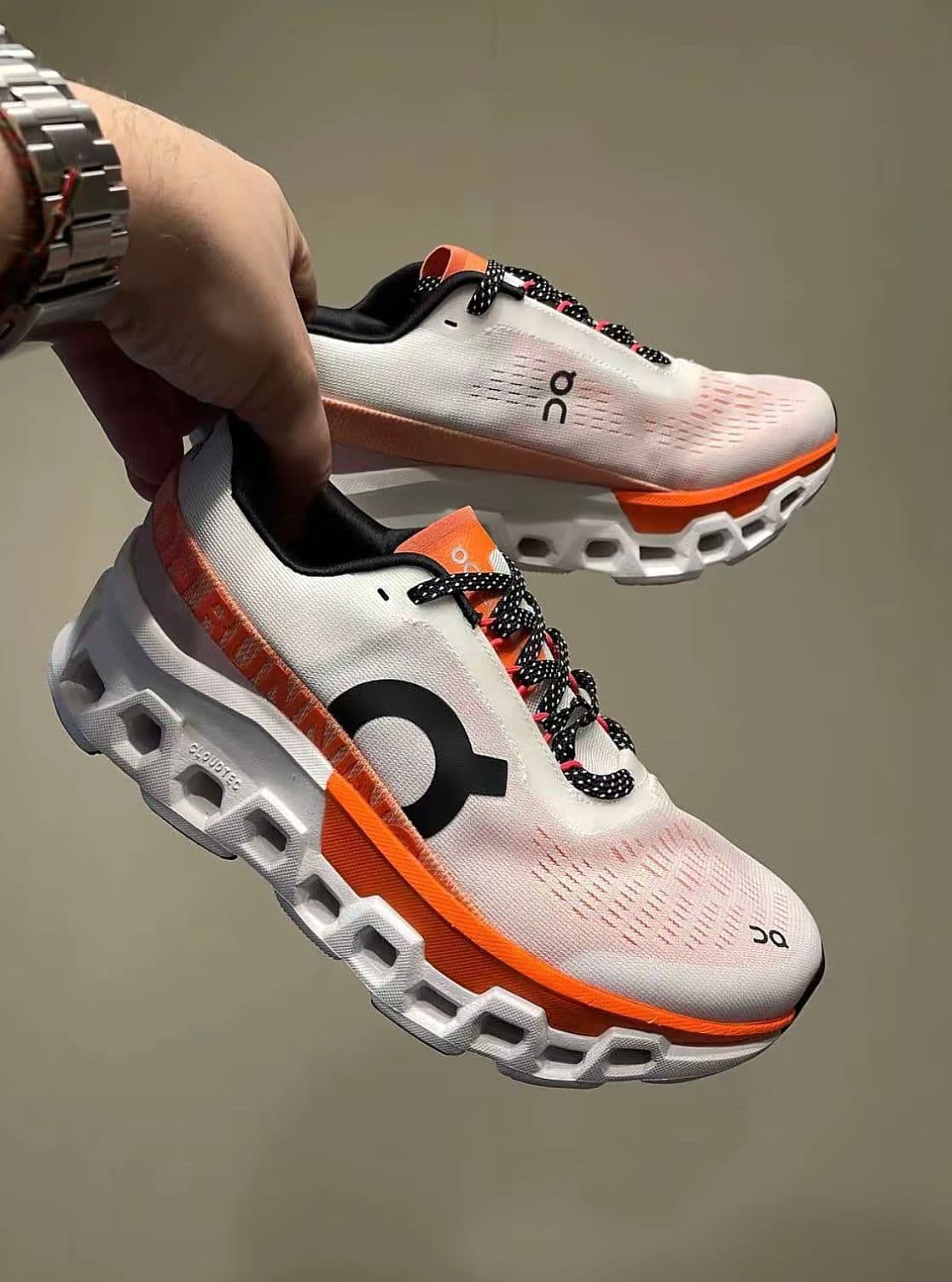 0ba0c3f9-58a7-4c41-93be-b53693f39d02 | AE JERSEY White On running shoes with orange accents and black-and-white laces being held mid-air against a neutral background, showcasing the design.