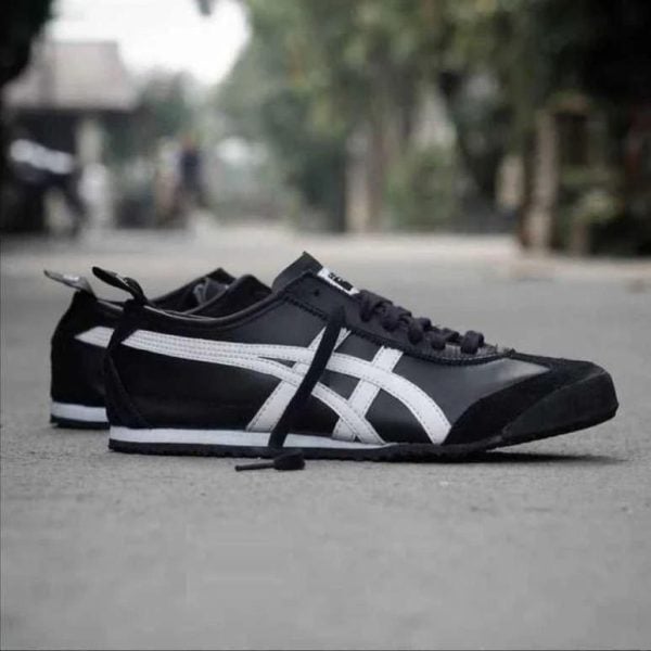 Pair of black sneakers with white stripes lying on a street surface, one shoe slightly ahead of the other on the ground.