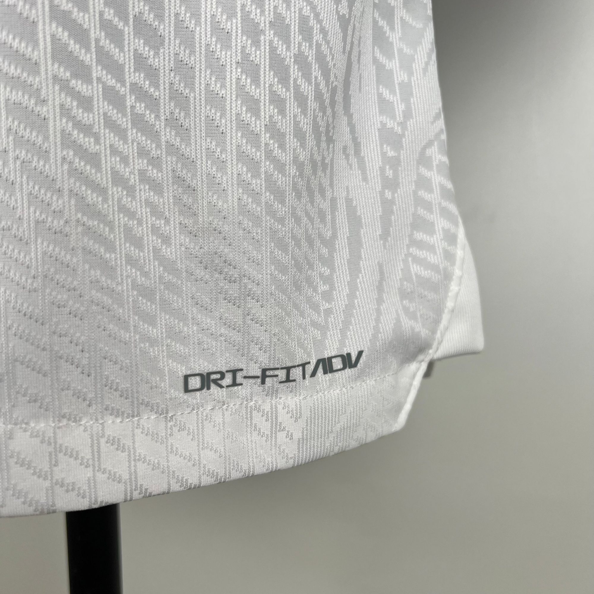 0d102d42_a3666112-f1c4-4dea-a83c-45c654b5b05f | AE JERSEY Close-up of white patterned chair fabric with the text DRI-FIT/ADV and part of a black chair leg visible to the left.