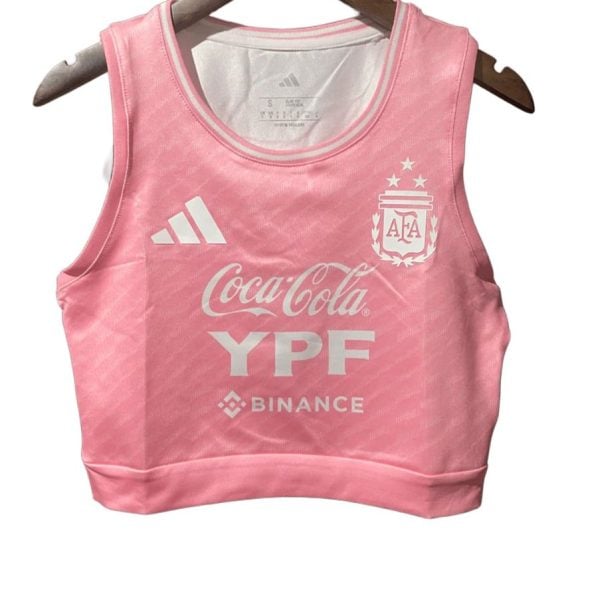 Pink cropped athletic top with Adidas logo, Argentina crest, and sponsor logos (Coca-Cola, YPF, Binance) on chest, hanging on a wooden hanger in front of a white background.