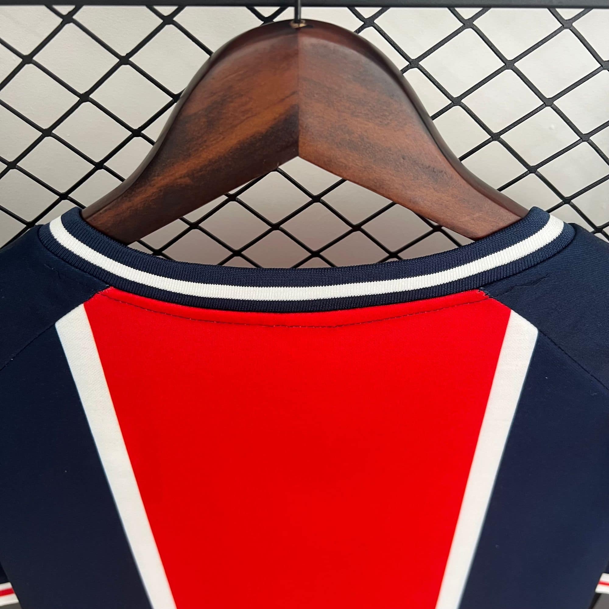 0ea03ed3_8e48ea0e-4fd0-4999-9c34-106de996fc85 | AE JERSEY Red athletic jersey with white and navy trim hung on a wooden hanger in front of a chain-link fence in the background.
