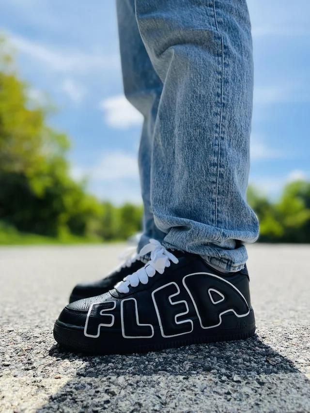 0f6455d8-a90a-44a0-99dc-37b9391c6acd | AE JERSEY Close-up of a person wearing black sneakers with white laces and large white outlined letters forming 'FLEA' on the side, standing on a paved road with jeans cuffs.