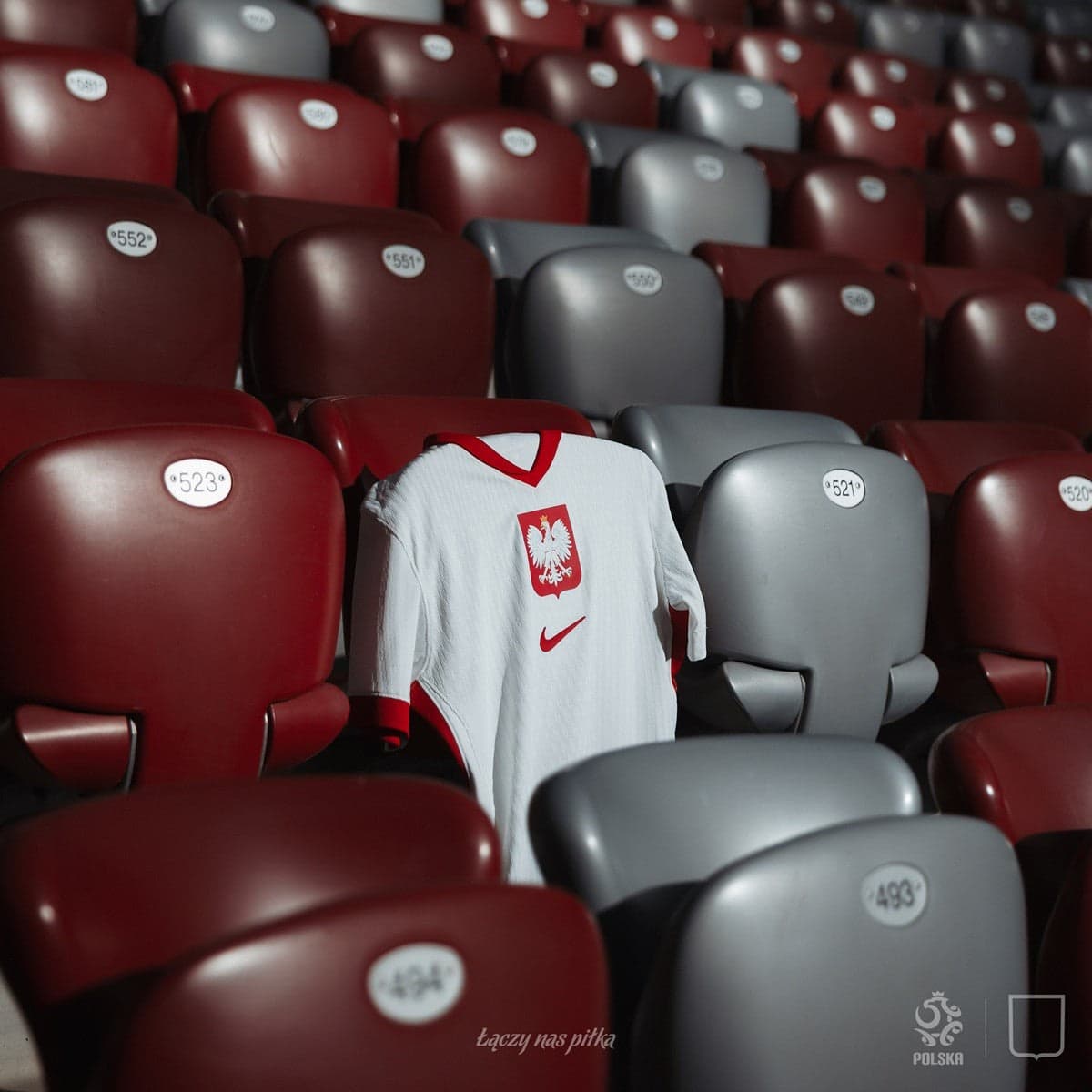 0twxlDVIp0Af48u | AE JERSEY White Poland national soccer jersey draped over red stadium seats in an empty arena with eagle crest and red trim