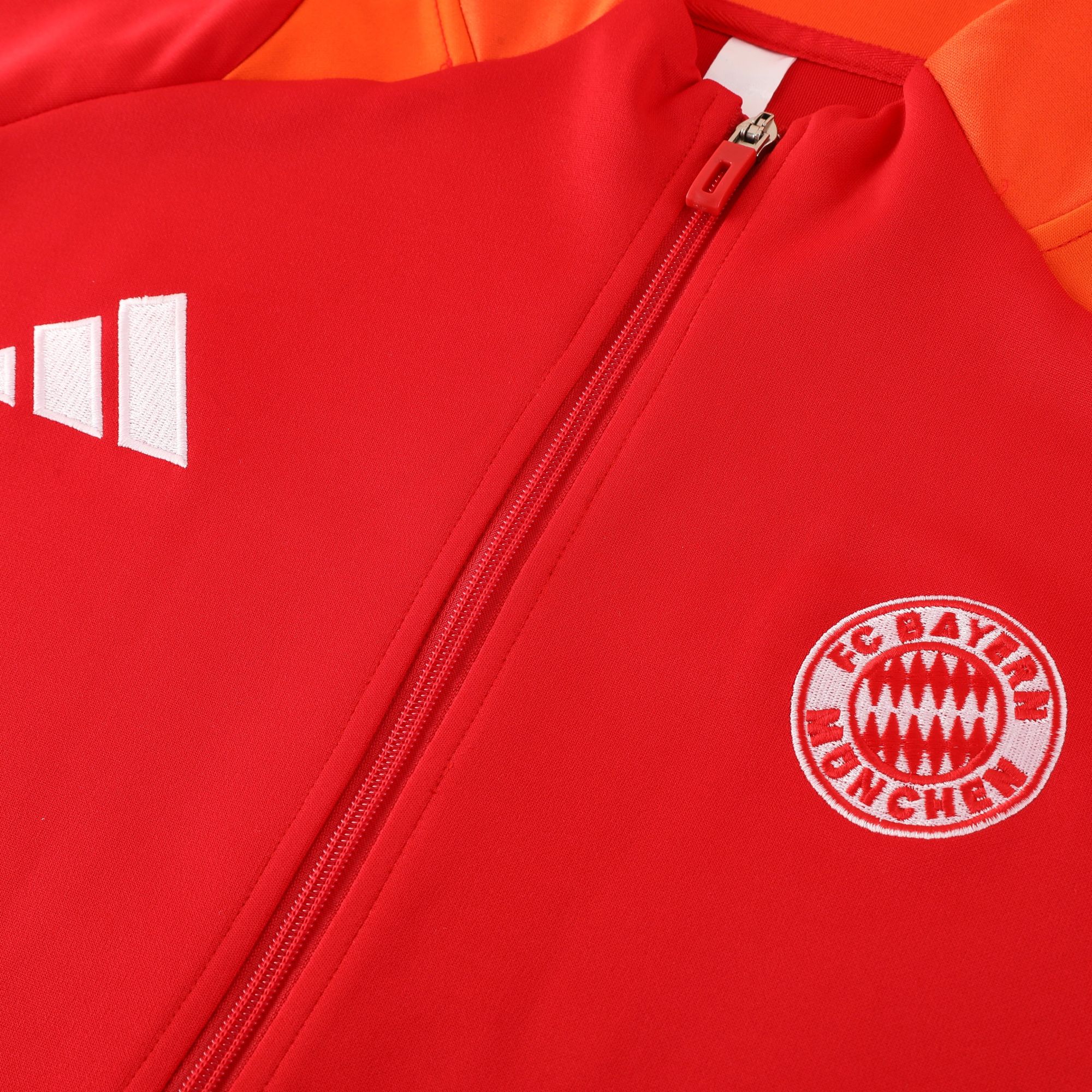 10fc3e0e | AE JERSEY Close-up of a red sports jacket zipper with white stitched patches and a Bayern Munich crest visible in the bottom-right corner.