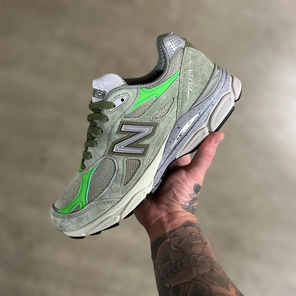 1179fdf3-12cb-4599-9352-78f3fb31e178_f6e39a38-7d95-47ba-9eb7-d98f793df827 | AE JERSEY Gray-green New Balance sneaker held in a tattooed hand, showing side logo and green accents.