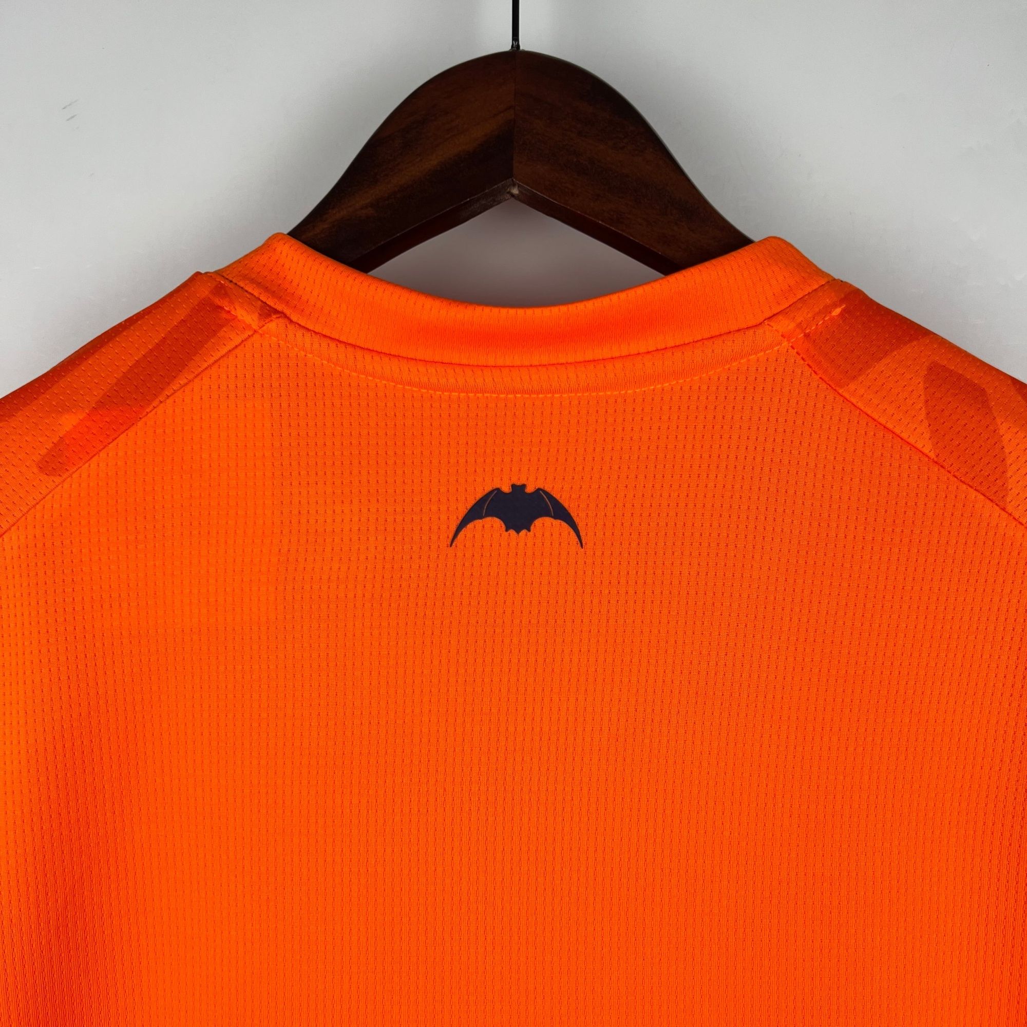 137cb007_16555ed2-0cb9-48b4-b5aa-cced7d8f5781 | AE JERSEY Bright orange textured polo shirt hanging on a wooden hanger, viewed from the back against a gray wall.