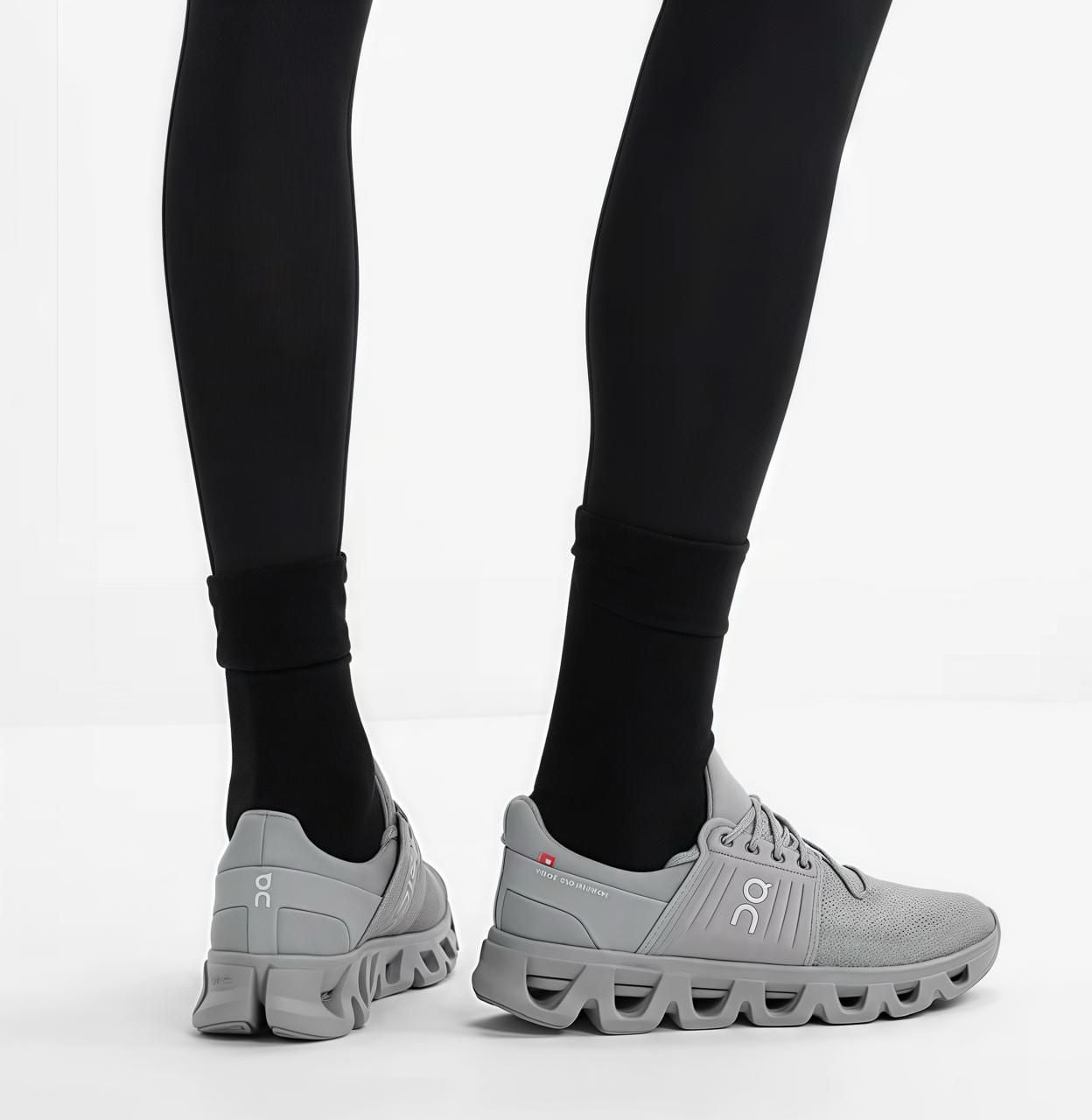 165e1cbe-23bb-4e7a-9726-8bc24c9faf2b | AE JERSEY Close-up of legs in black leggings wearing light gray On running shoes with chunky, wavy soles on a white floor.