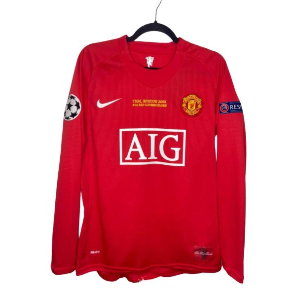 Red Manchester United long-sleeve jersey on a hanger showing the club crest, white Nike swoosh, and large AIG sponsor logo on the front; UEFA patch on the left sleeve and small text/details visible at the bottom hem.