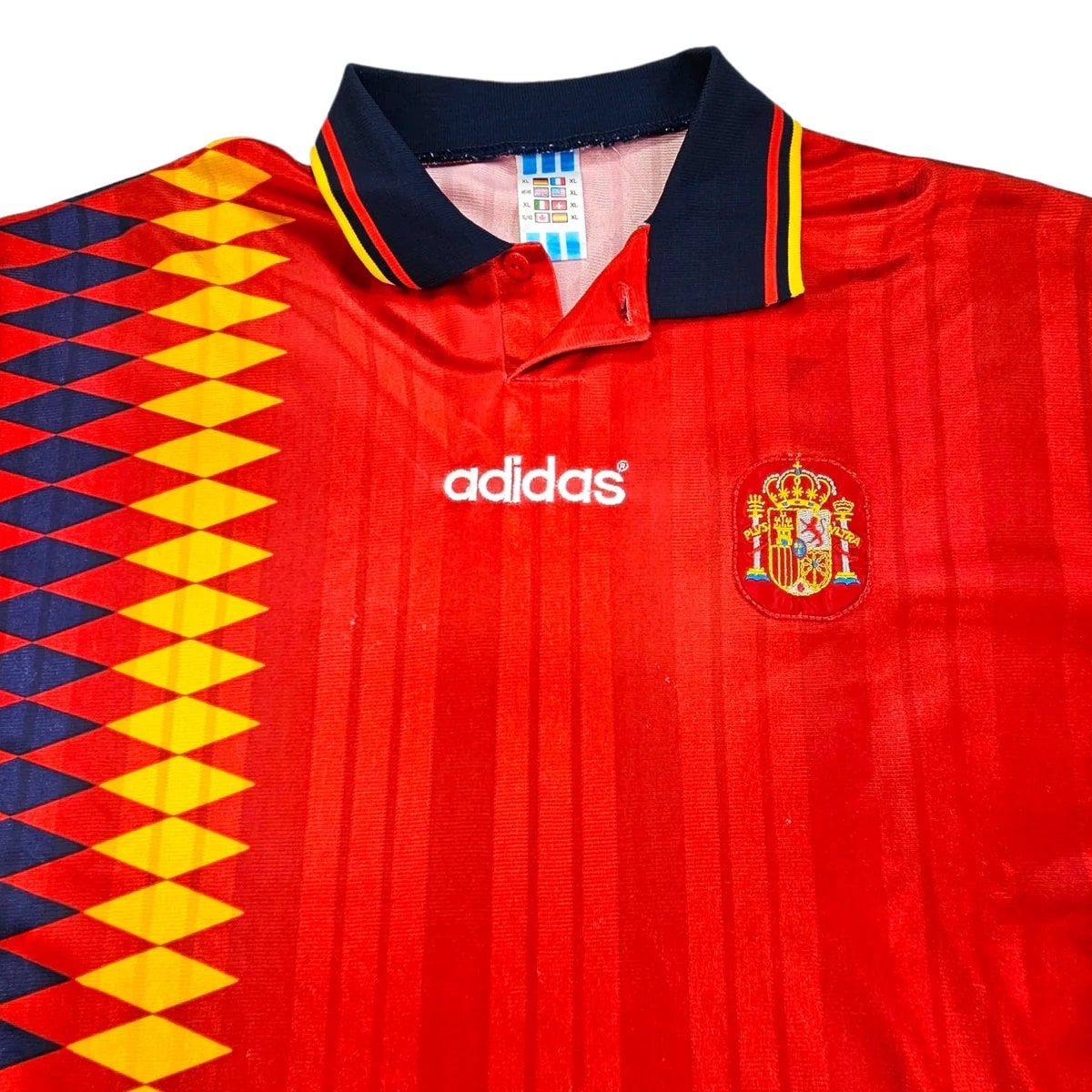 199496-spain-home-football-shirt-xl-adidas-ff202729-674014 | AE JERSEY Red Spain national football jersey by Adidas, featuring a dark navy collar with yellow and red trim and a geometric yellow-blue pattern on the left panel, plus the Spain crest on the chest.