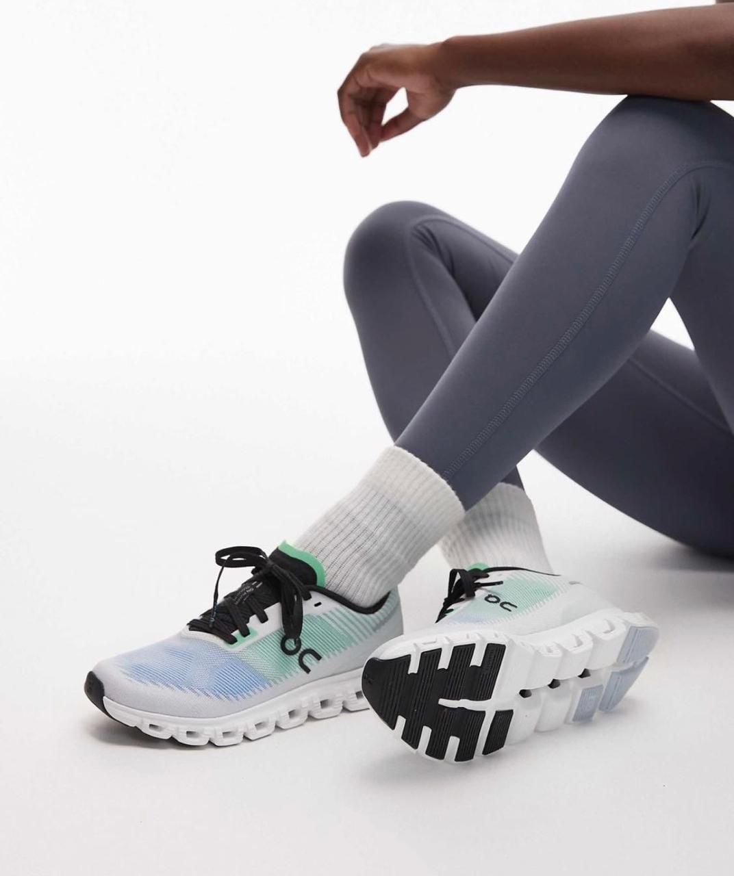 19a4f039-35d8-4441-8970-6e2464cac4de | AE JERSEY Close-up of white and teal running shoes with chunky sole, worn with gray leggings and white socks, on a white floor.