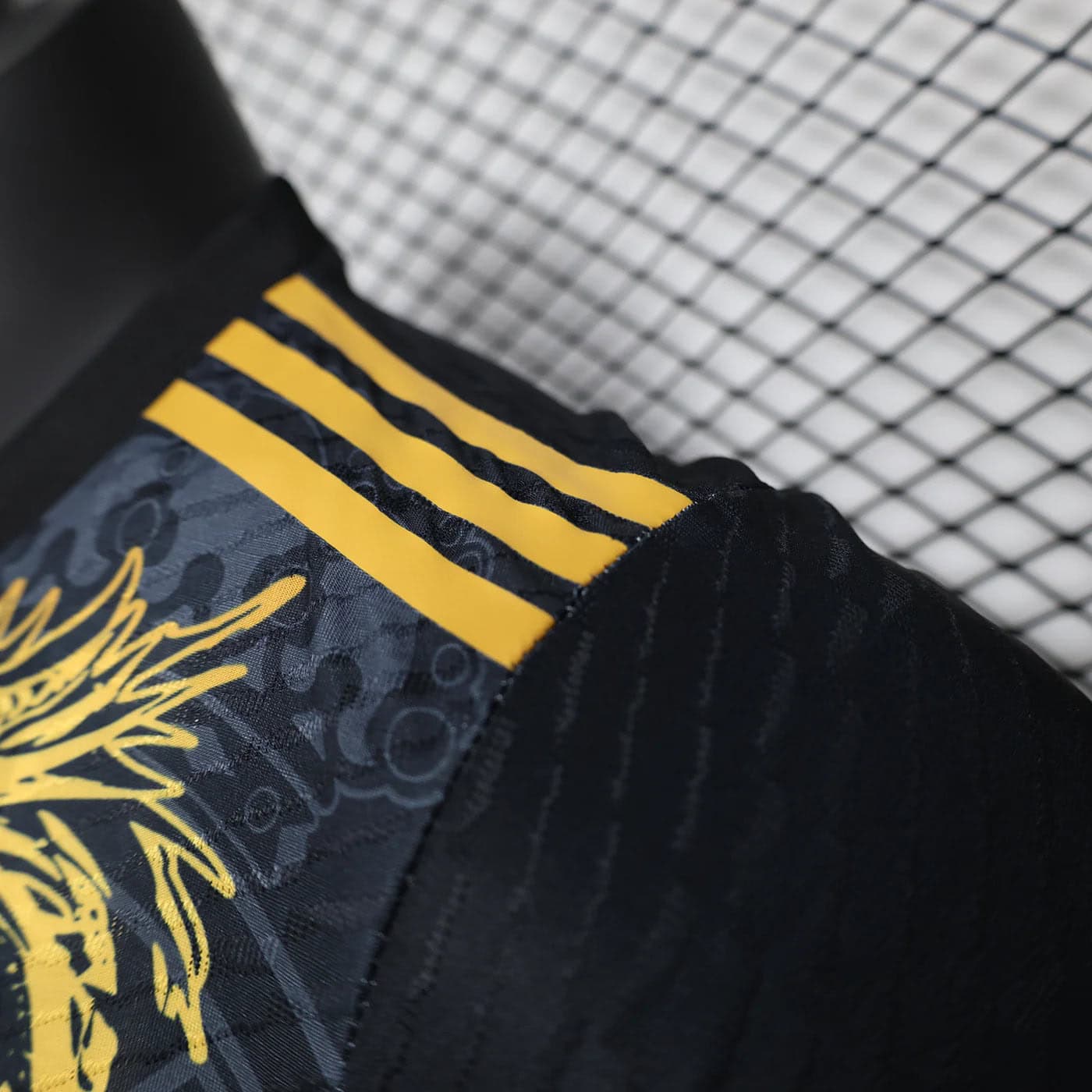 1a86dc83 | AE JERSEY Close-up of dark fabric with three yellow stripes and a gold graphic pattern, likely athletic wear detail, against a chain-link fence background.