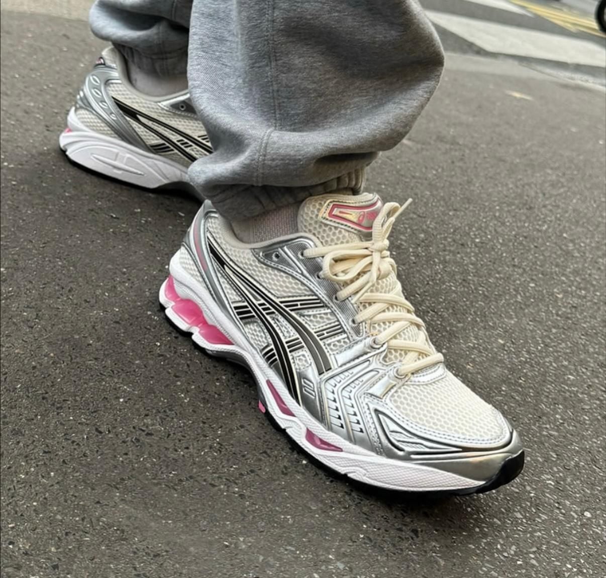 1e381fc7-c917-409e-b456-f01d19f88fd6 | AE JERSEY Close-up of a person wearing light gray sweatpants and silver-white running shoes with black stripes and pink accents, standing on a paved street.