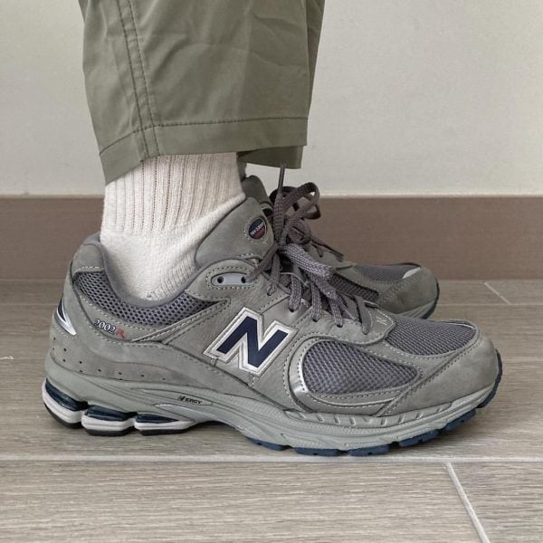 Grey New Balance athletic sneaker with white sock and olive pants, shown from the side on a tiled floor, 2002R model detail visible