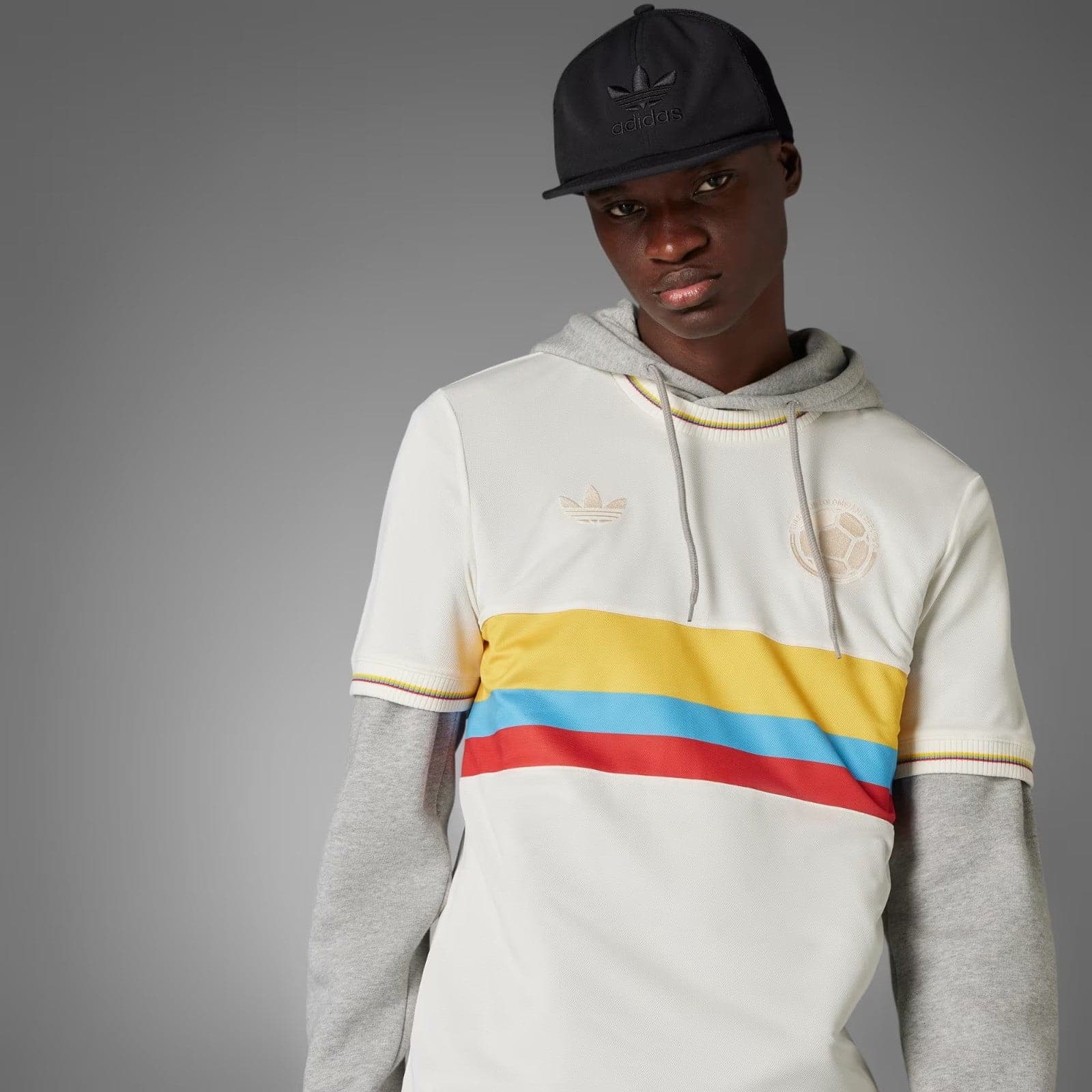 1sjy4mYQrdCrM5u | AE JERSEY Model wearing a white color-block hoodie with gray sleeves and a multicolor stripe, paired with a black Adidas cap against a gray studio backdrop.