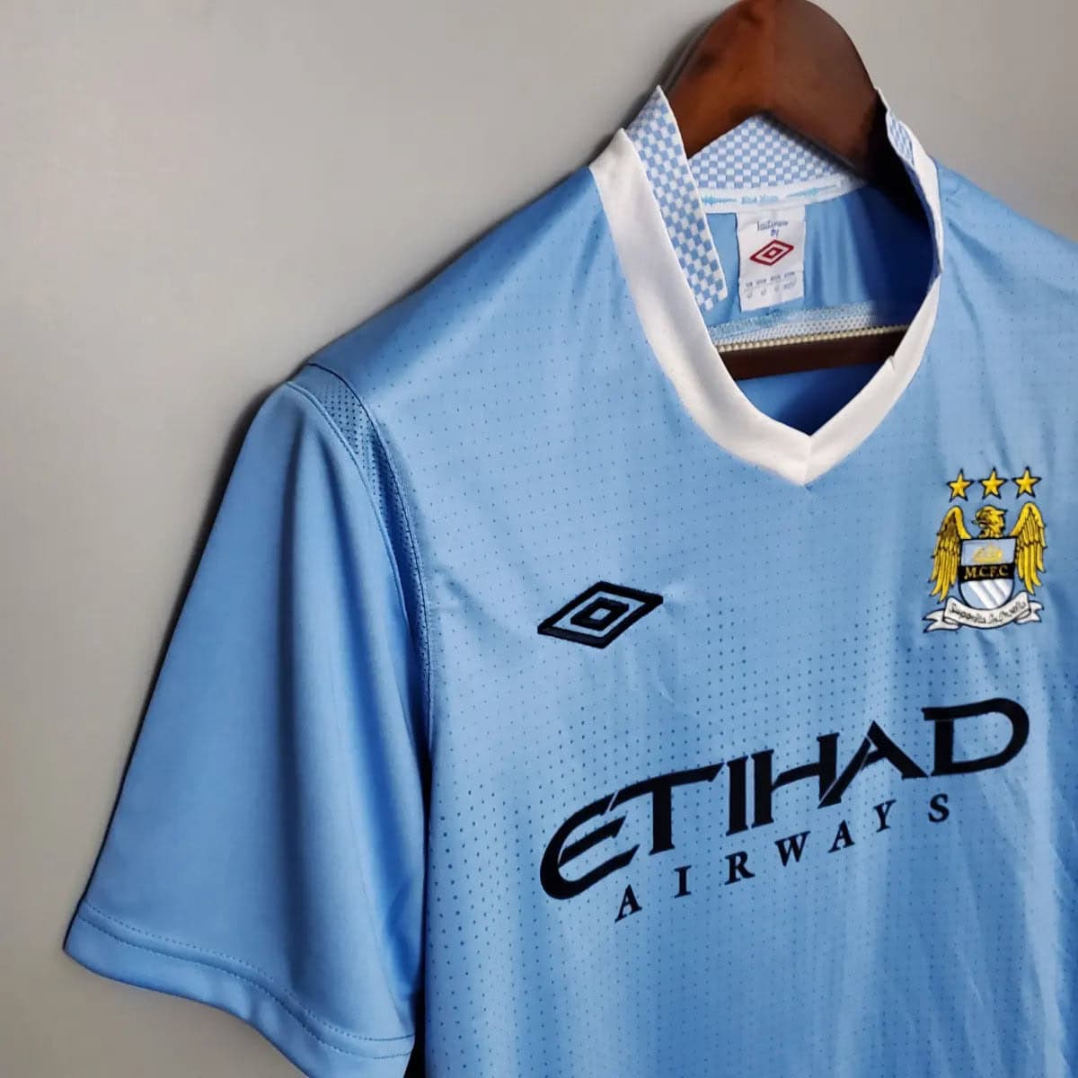 2012-Manchester-City-Retro-Home-Short-Sleeve-Jersey5 | AE JERSEY Light blue Manchester City football jersey on a wooden hanger, white collar and sponsor text Etihad Airways.