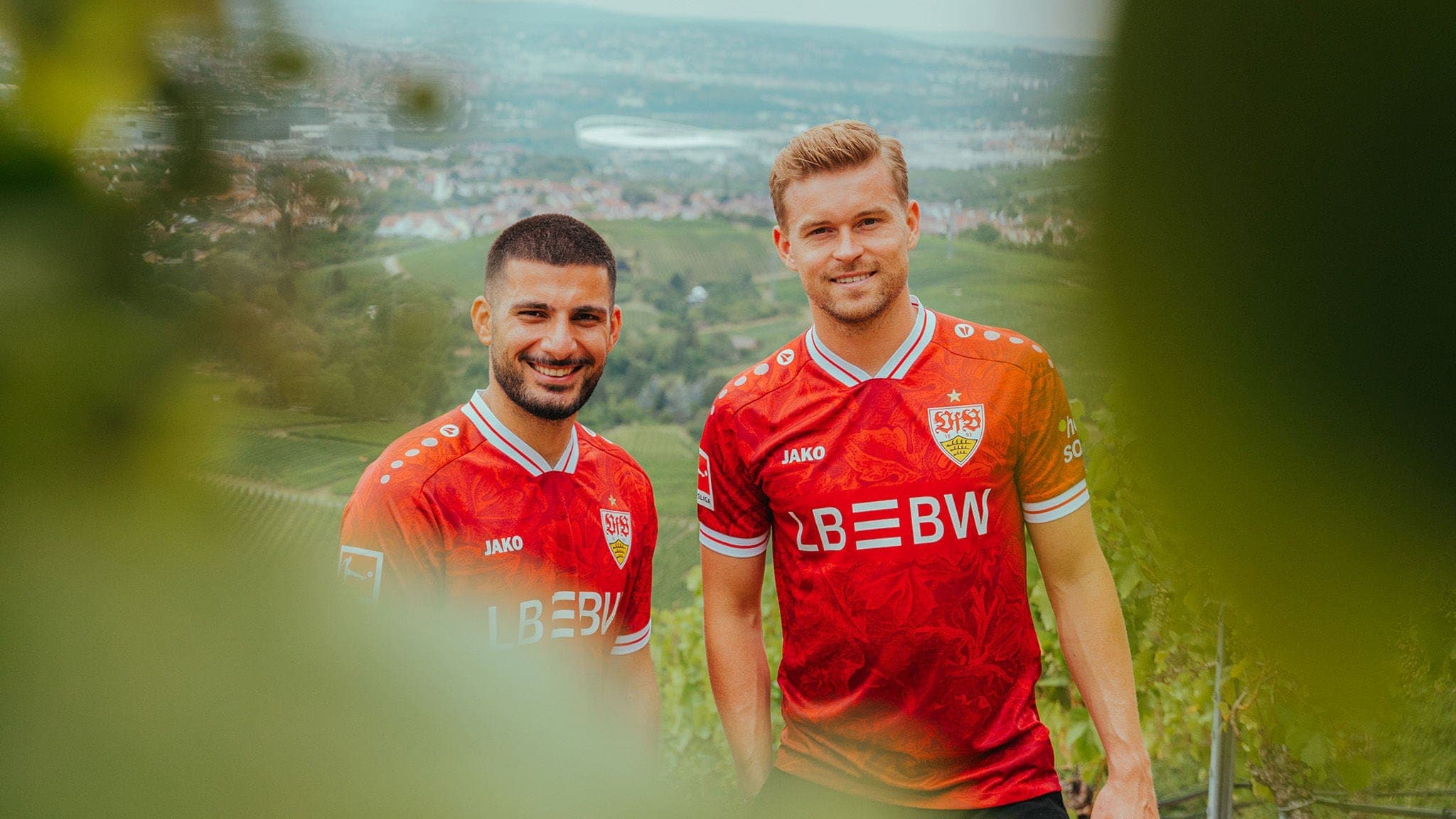 2025-2026-VfB_Auswaertstrikot-02_Rot_Spieler_00000_x_0x0_8405aff0-5217-4329-859b-2425681e2170 | AE JERSEY Two male soccer players in red jerseys smile for the camera against a hillside town backdrop with blurred greenery in the foreground.