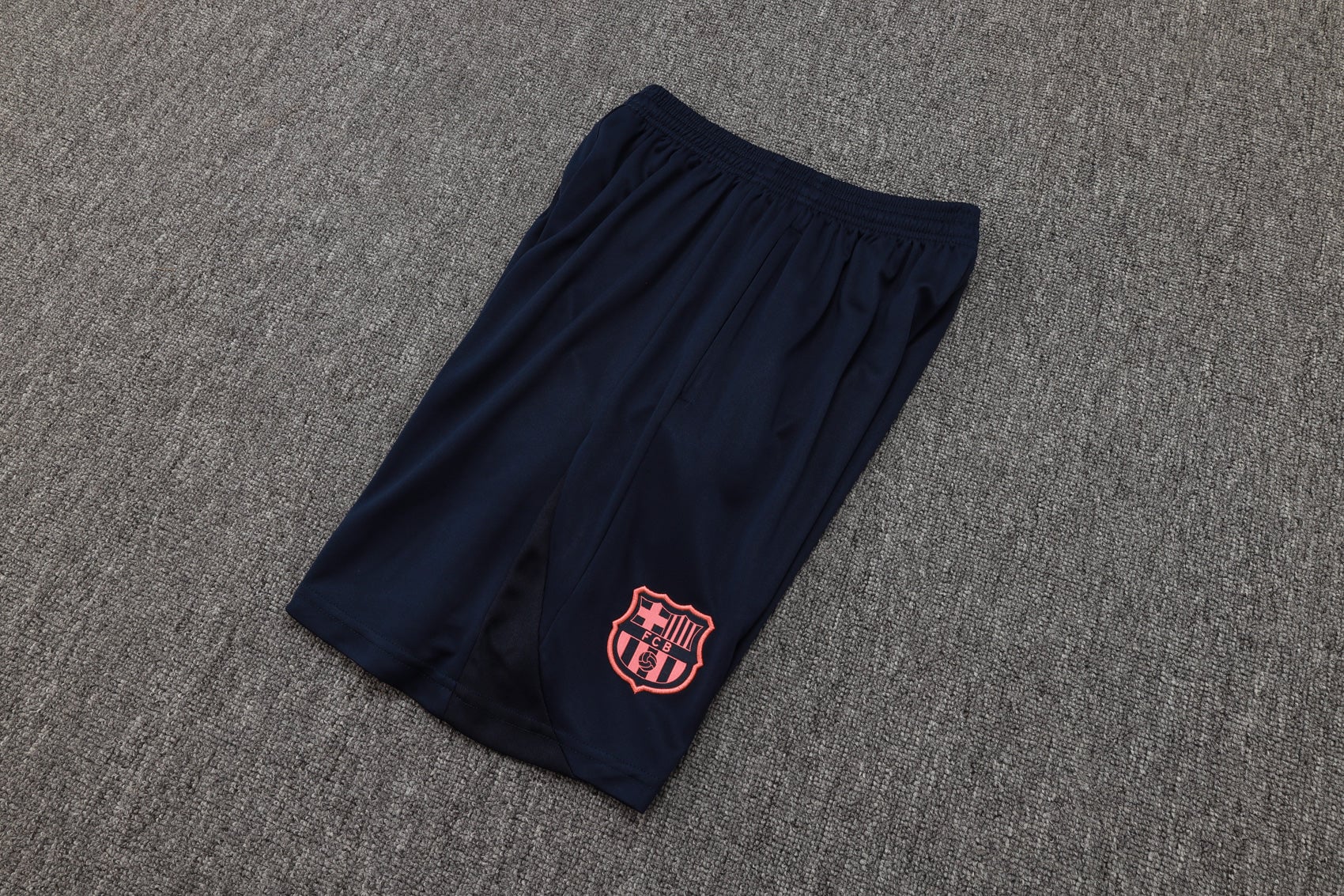 202c12c2 | AE JERSEY Navy athletic shorts with a pink FC Barcelona crest on the lower leg, laid on a gray carpeted floor.