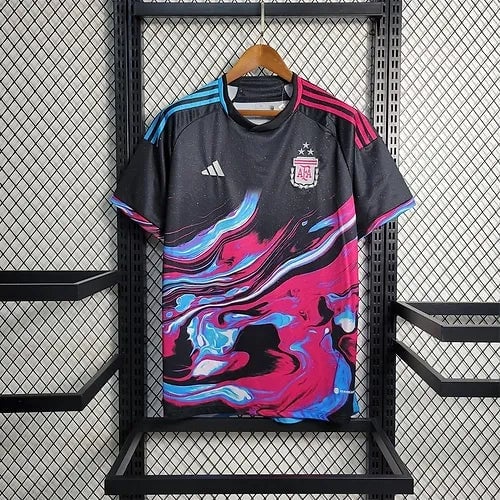 20903b_af1ad6f97275438f9ddc7ce4ad0f0613_mv2 | AE JERSEY Black Adidas Argentina soccer jersey with a bold pink, blue, and white swirl design, hanging on a wooden hanger against a metal grid display.
