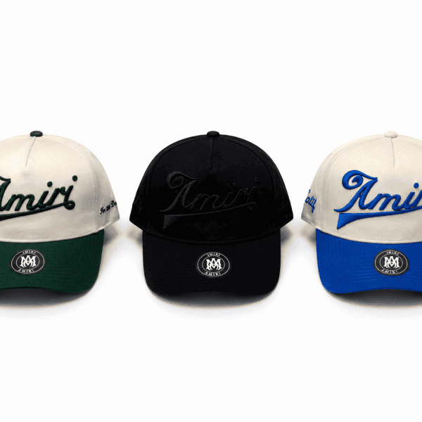 Three Amiri baseball caps: cream with green brim, black, and cream with blue brim; 'Amiri' script on the front edge and a circular patch on the bill area.