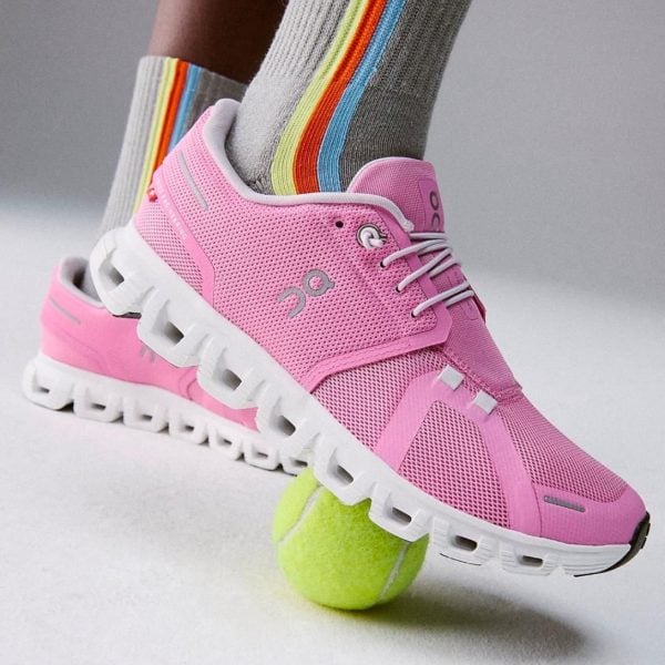 Pink athletic running shoe stepping on a bright tennis ball, with striped rainbow socks visible behind it.