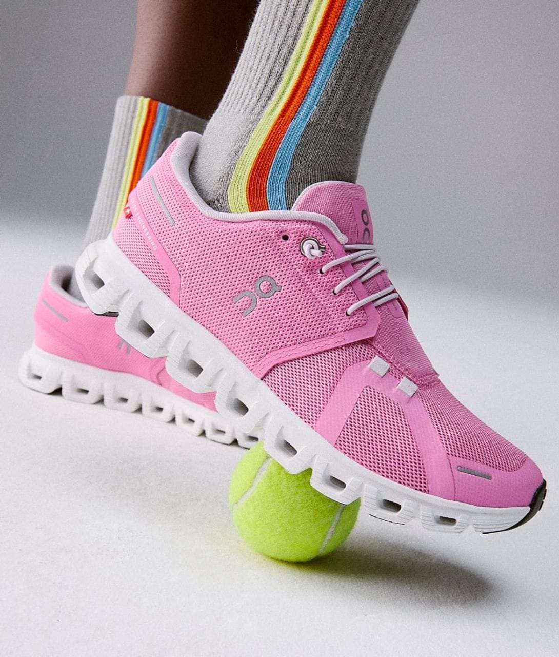 27a68a48-51d6-498c-a535-b993a1f614d0 | AE JERSEY Pink athletic running shoe stepping on a bright tennis ball, with striped rainbow socks visible behind it.