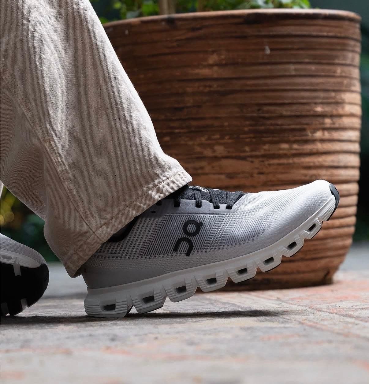 28dd7f84-b6c7-469b-953e-4384fd6cafc8 | AE JERSEY Close-up of a gray running shoe on a person’s foot, beige pants, stepping on a paved surface with a wooden planter in the background.