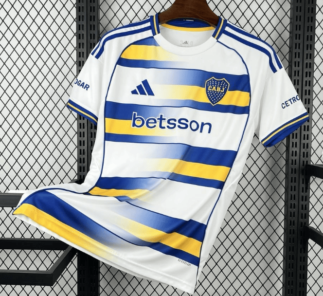 2_05ce222e-07ad-4328-95a1-2ede47d6b72c | AE JERSEY White soccer jersey with blue and yellow diagonal stripes, CABJ crest, Betsson sponsor, hanging on a wooden hanger against a metal grid background.