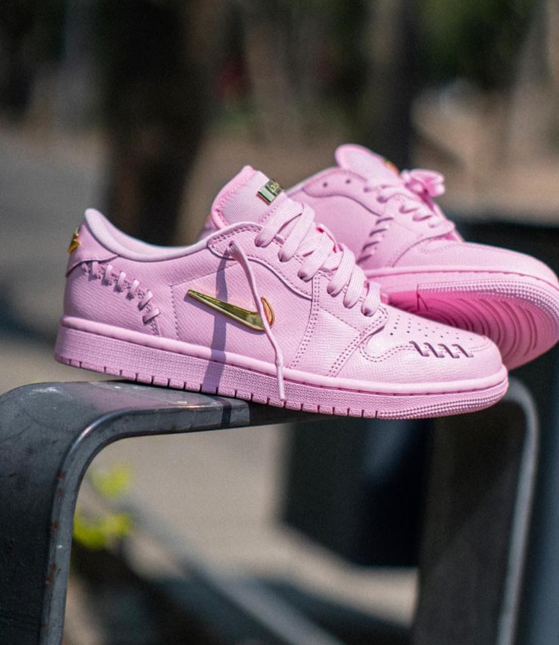 2cbc93fa-1f00-4719-9029-a825721ef694 | AE JERSEY Pair of pink Nike sneakers with a gold swoosh resting on a metal railing outdoors.