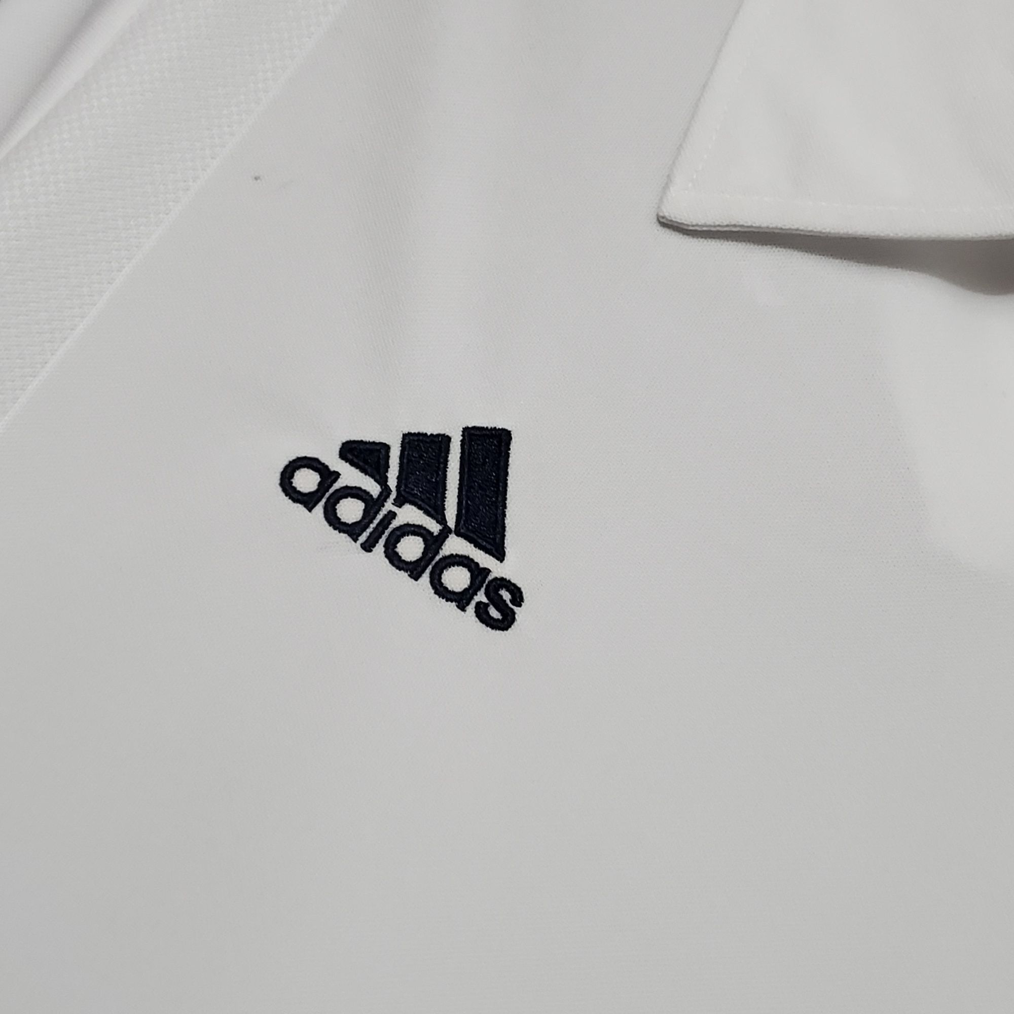 30ba54e8_c41f6699-8f90-445c-8458-9e2df11e83f9 | AE JERSEY Close-up of white fabric with the Adidas logo partially visible in black letters, cropped at the bottom.