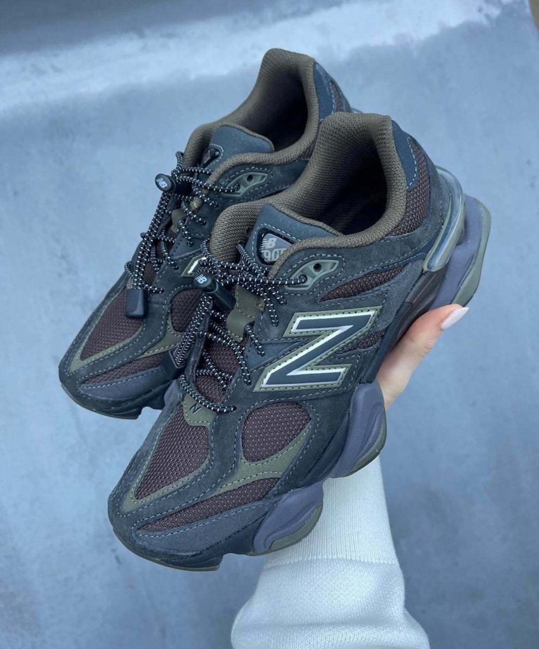 31027769-4664-4858-a6e4-8e360767e56f | AE JERSEY Dark gray New Balance 901 trail sneakers with brown overlays, held in a hand against a gray background, showing the 'N' logo and rugged sole