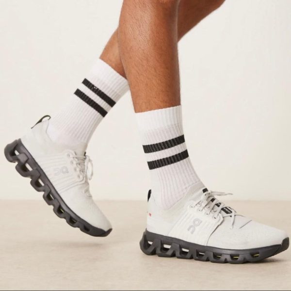 White athletic sneakers with a distinctive black segmented sole, worn with white crew socks featuring two black stripes at the top edge.