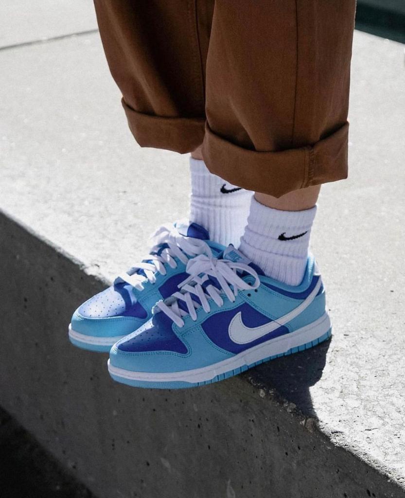 3500ee53-de16-4e66-878b-e23811eaff7a_e1b3233c-7bad-4b8b-8088-a413ea1c826c | AE JERSEY Light blue Nike sneakers with white laces on white socks and rolled brown pants, standing on a concrete curb.
