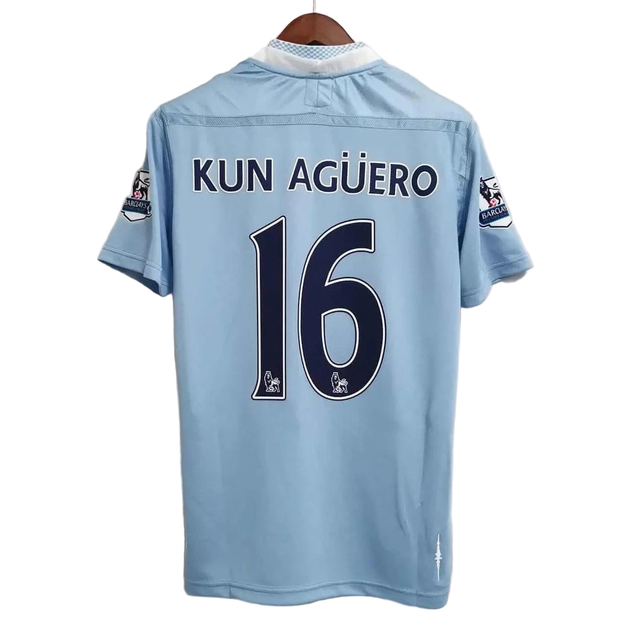 35d2e41c-1-e1662940375495 | AE JERSEY Back view of a light‑blue Manchester City jersey with the name KUN AGÜERO and the number 16 printed on the back, hanging on a wooden hanger on a white background