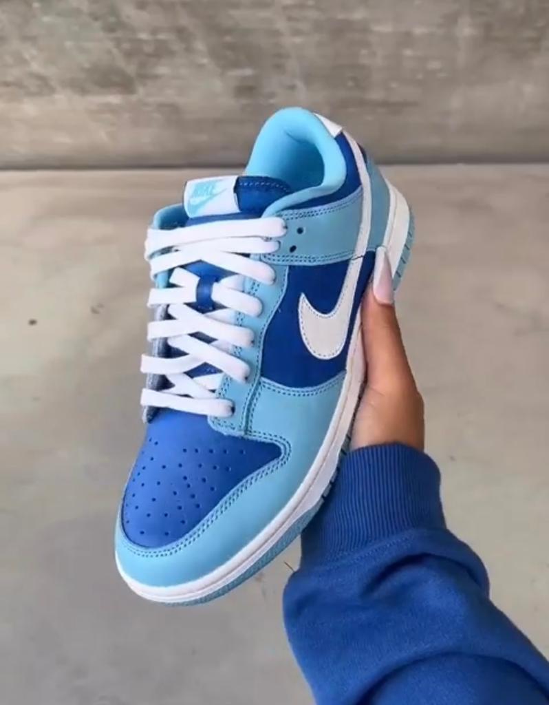 3897ef3a-5020-49de-bae9-186fe23e4951_f4208658-d07f-4dc6-95ae-ba40b22a3f3b | AE JERSEY Blue Nike sneaker with white laces held in a hand, showing light and dark blue panels against a concrete background.