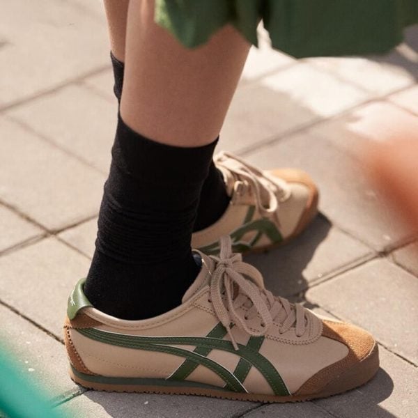 Close-up of beige sneakers with green stripes and brown toe caps worn with black socks on a paved sidewalk.