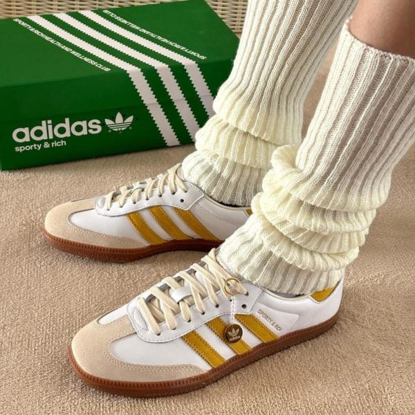 White Adidas sneakers with yellow stripes and cream suede toe caps, paired with thick ribbed cream socks, beside a green Adidas shoebox on carpeted floor.