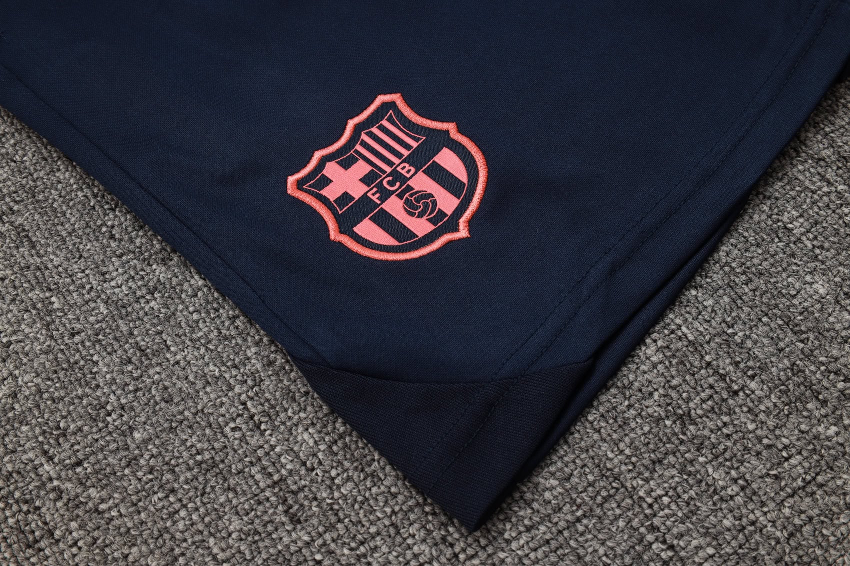 3f9cd96c | AE JERSEY Navy fabric with a pink-embroidered FC Barcelona crest on the folded edge, resting on a gray carpeted surface.