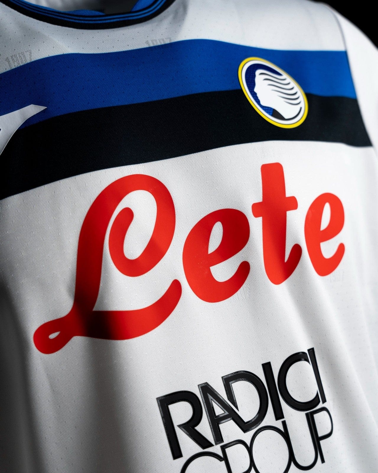 3v9Pw70CnxAOU9Y | AE JERSEY Close-up of a white jersey with red 'Leite' sponsor and a circular crest on a blue stripe near the chest area.