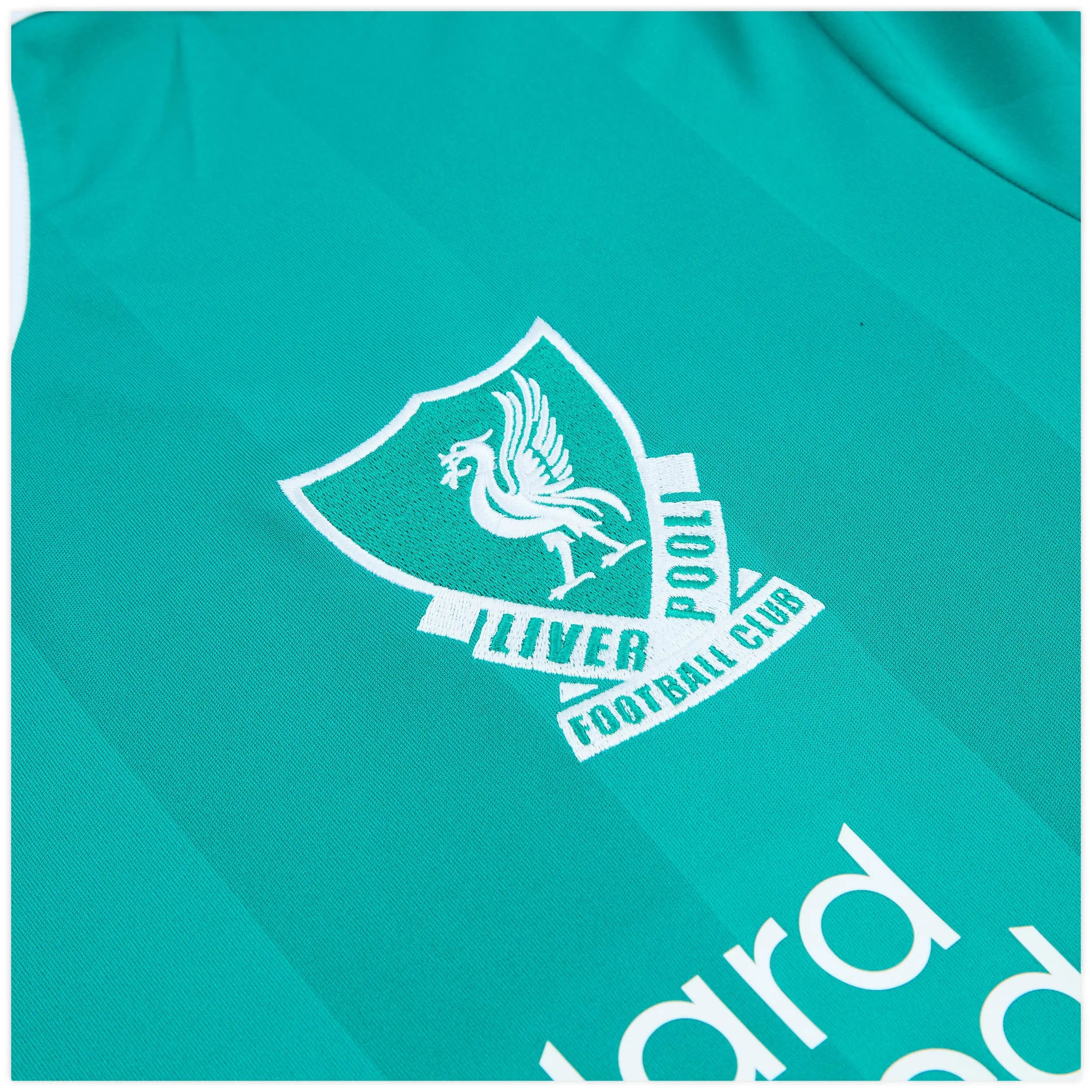 4068809557572-3_kxmfrjuhqmgeeuwp | AE JERSEY Teal fabric with a white embroidered shield logo and partial text, likely a sports club crest. The design is diagonally cropped across the image.