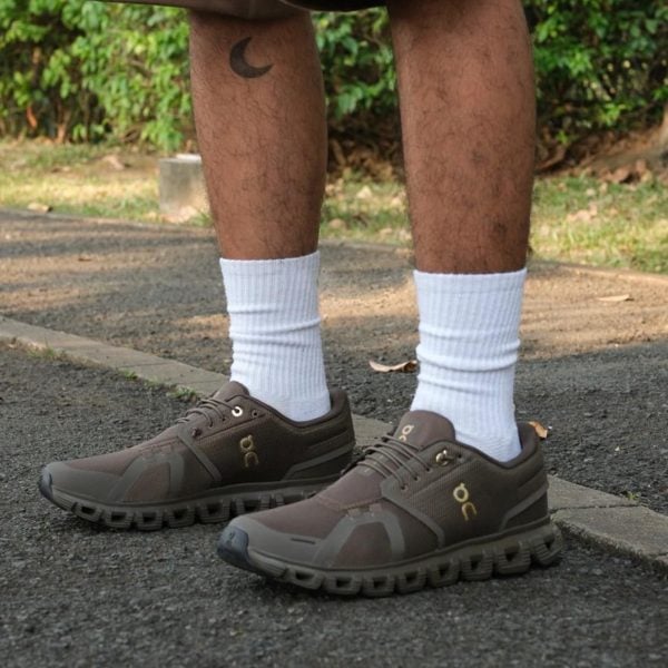 Lower legs wearing brown athletic shoes with white socks, standing on a paved path; crescent tattoo on the left leg visible.