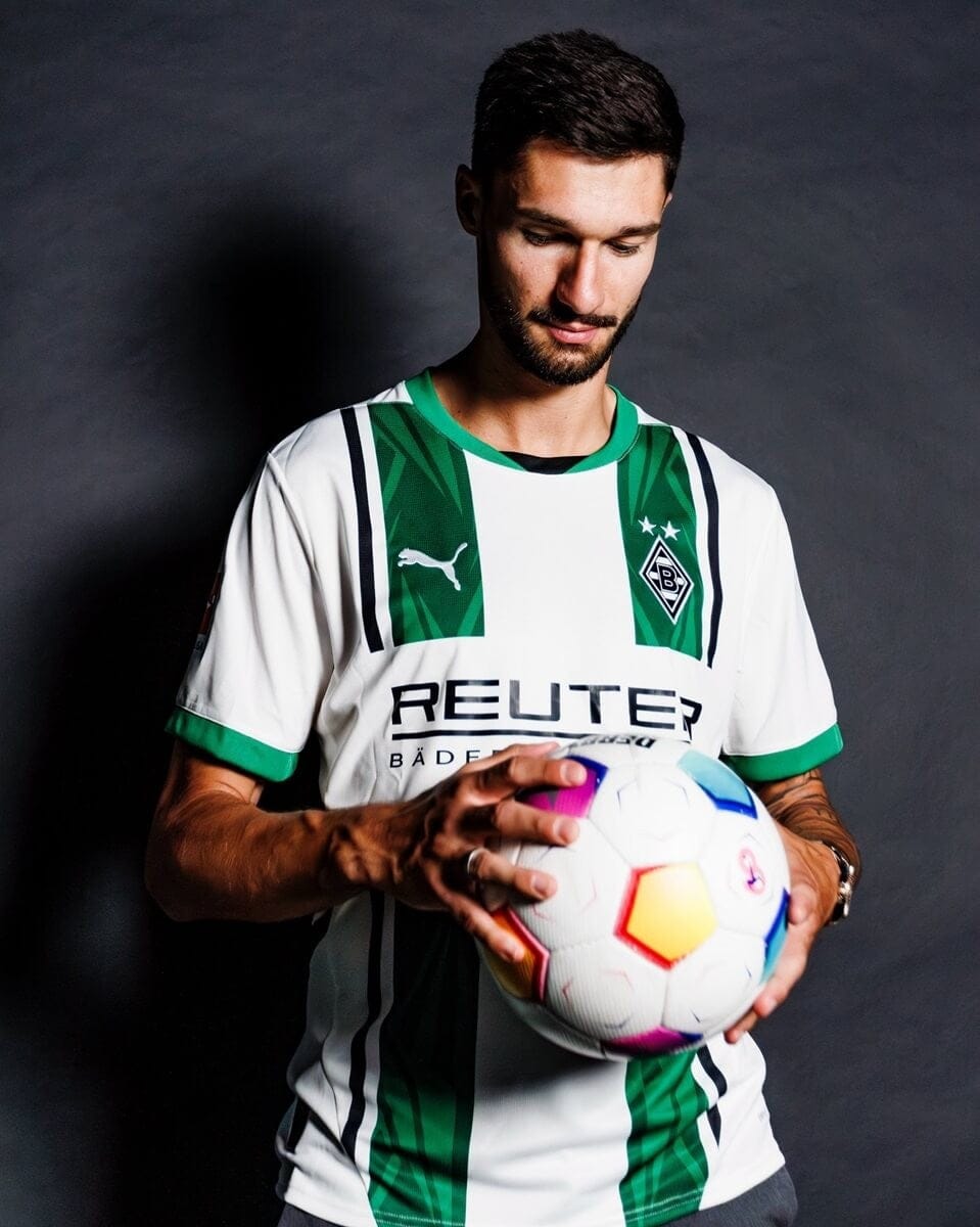 41KDrCKSfkGjBvh | AE JERSEY Male soccer player in a white and green jersey holds a colorful soccer ball, looking down at it against a dark backdrop.