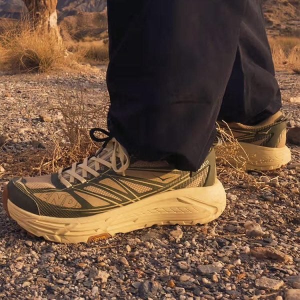 Close-up of olive-green hiking shoes on a rocky desert trail, with dark pants visible at the ankles.