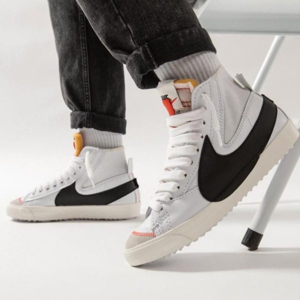 White high-top sneakers with a black side panel, worn with cuffed jeans in a bright studio setting.