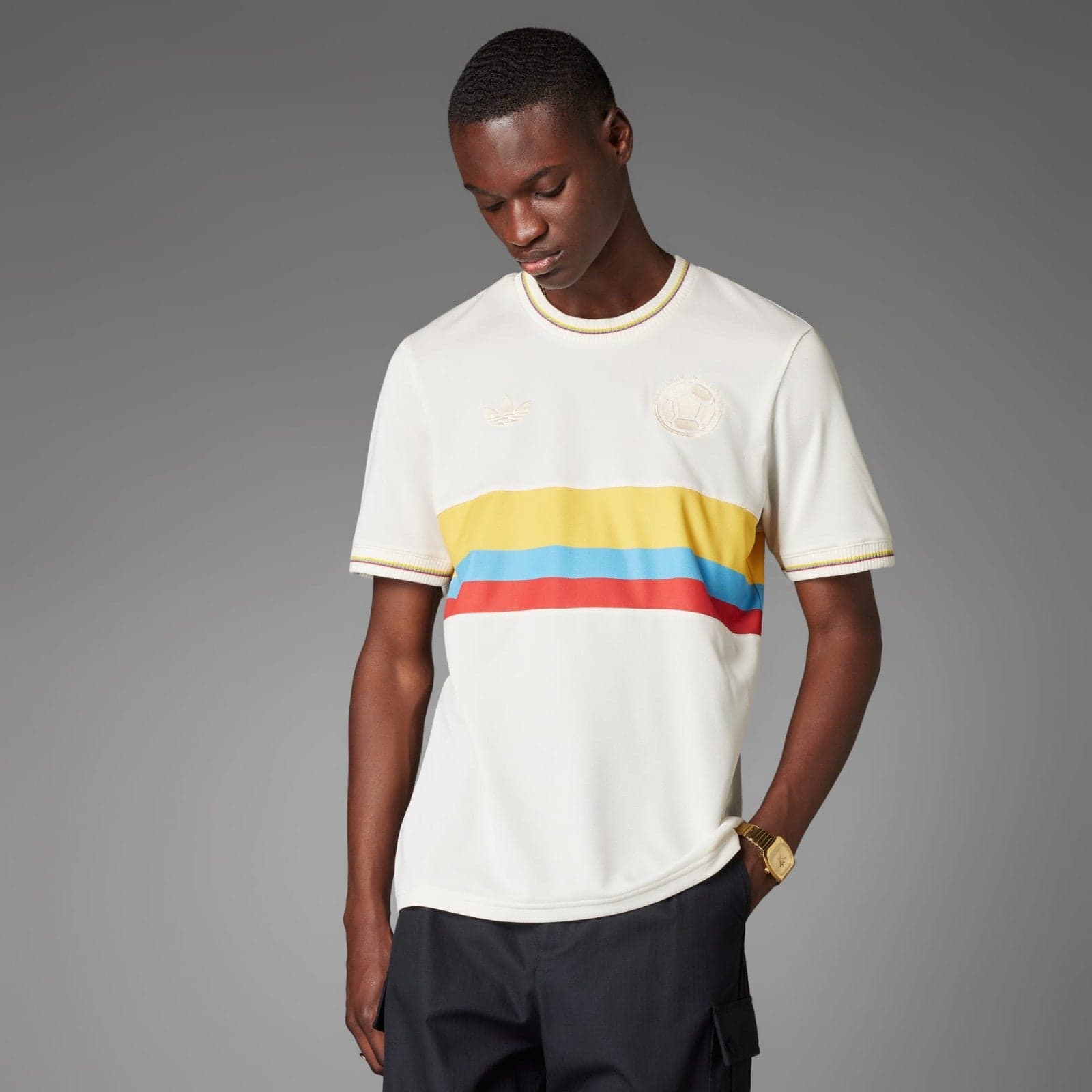 45IRTy0y9j8PfWa | AE JERSEY Young man wearing a white short-sleeve shirt with yellow, blue, and red horizontal stripes, looking down against a gray background.