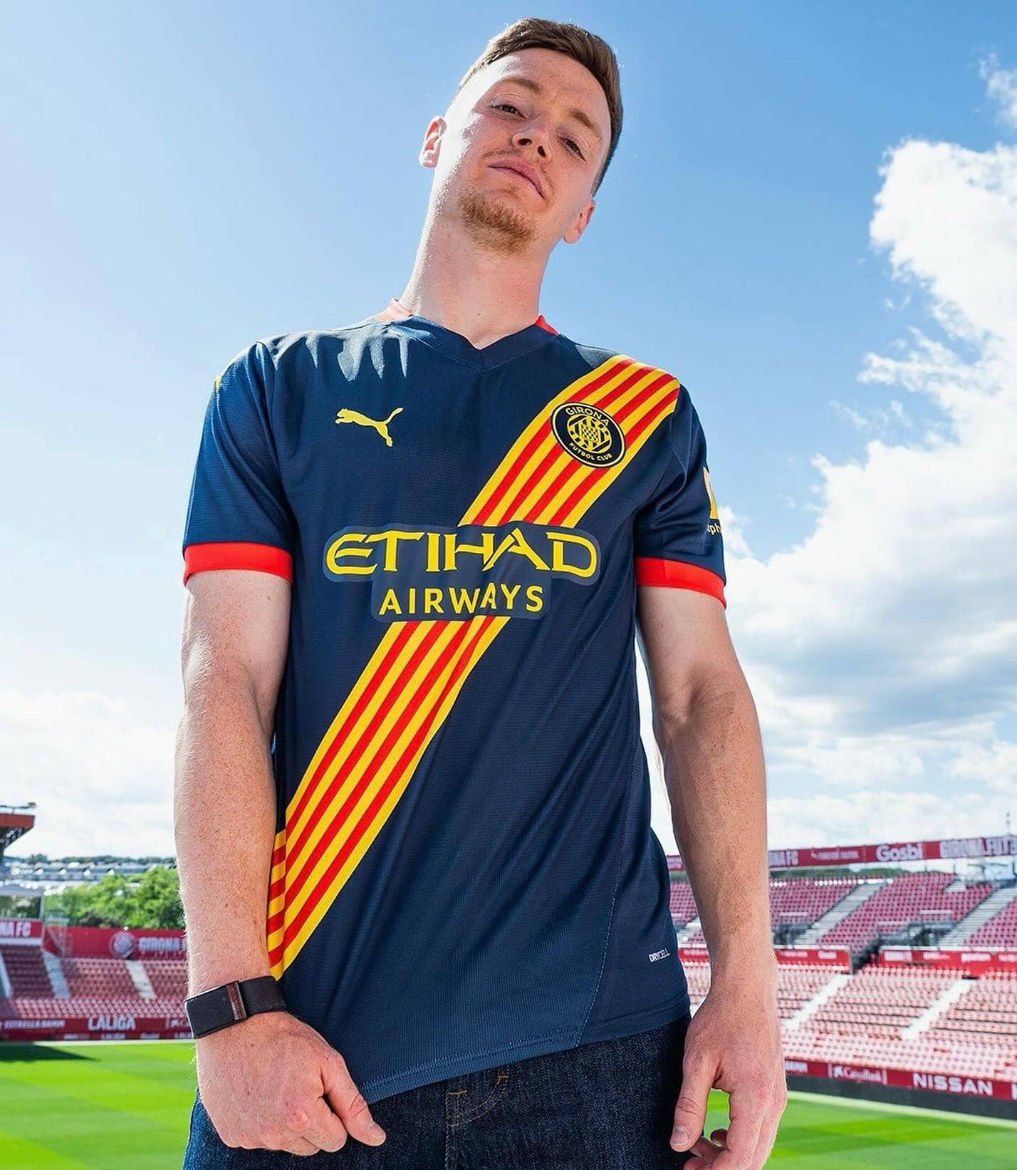 461505398_17940808712903197_5106101443471822670_n | AE JERSEY Young man wearing a blue Girona FC jersey with red-yellow diagonal stripe, standing in a stadium against a blue sky.