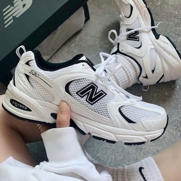 White New Balance 530 sneakers held in hand, showing mesh panels, white laces, and the NB logo.