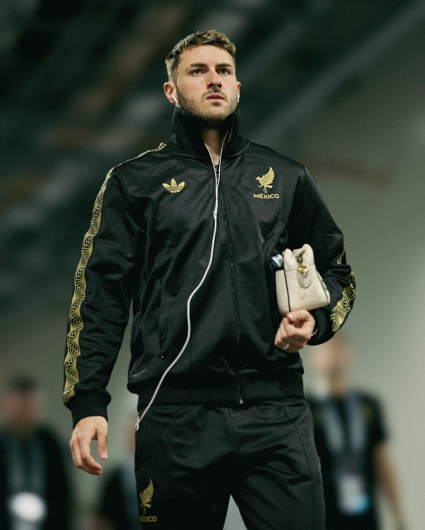 486040444_18496668016053657_4086695693259147033_n | AE JERSEY Athlete in a black Mexico jacket with gold emblem, wearing white earbuds, holding a small beige bag.